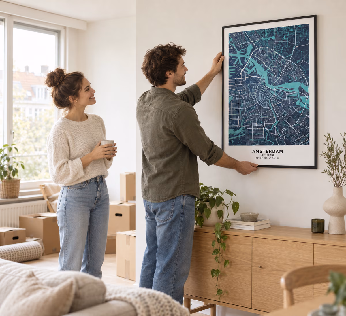 Een stel hangt een ingelijste stadskaart van Amsterdam op in hun nieuwe woonkamer, tussen de verhuisdozen